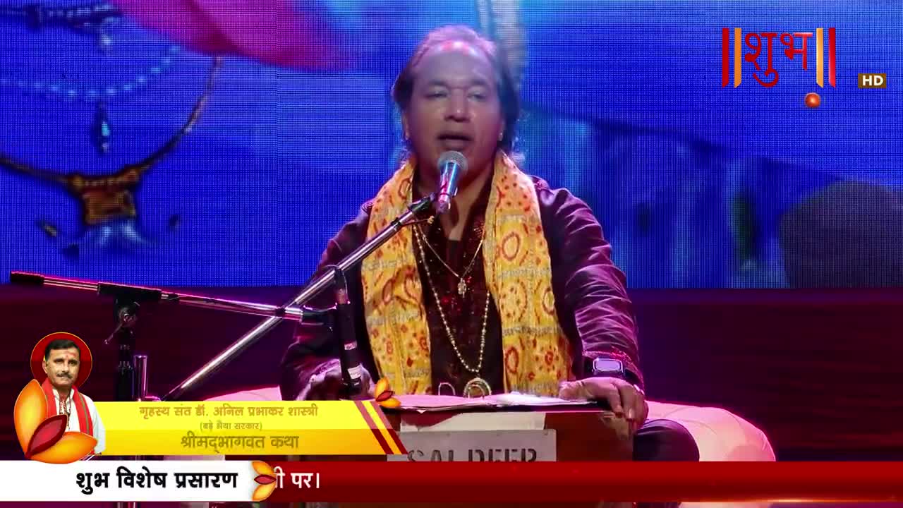 A man in a maroon outfit and a yellow scarf sings into a microphone, his hands resting on a harmonium. The Shubh TV logo appears in the upper right corner.
