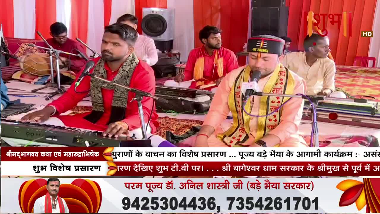 A man in a red kurta plays a keyboard while another in an orange robe sings into a microphone. The Shubh TV broadcast announces upcoming programs with a phone number displayed below.