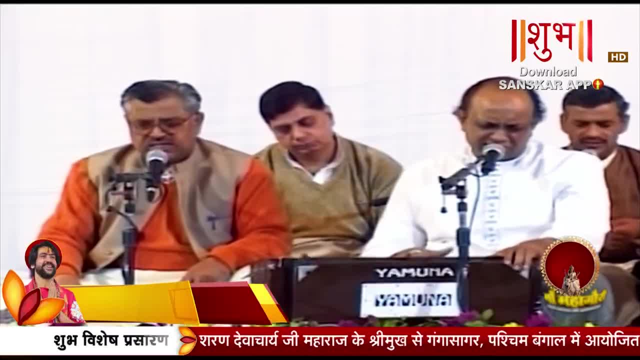 A man in a white kurta sings into a microphone, his fingers poised over a harmonium. Another man in an orange kurta sits to his left, also near a microphone.
