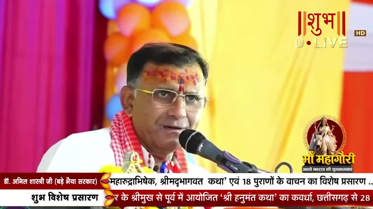 A man with a red tilak on his forehead speaks into a microphone. He wears glasses and a checkered scarf around his neck.