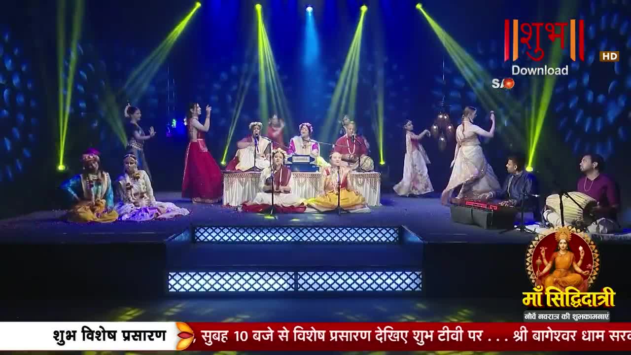 Dancers in traditional Indian attire move gracefully on a stage bathed in yellow spotlights. Musicians play instruments, including a harmonium and drums, as part of a performance for Shubh TV.