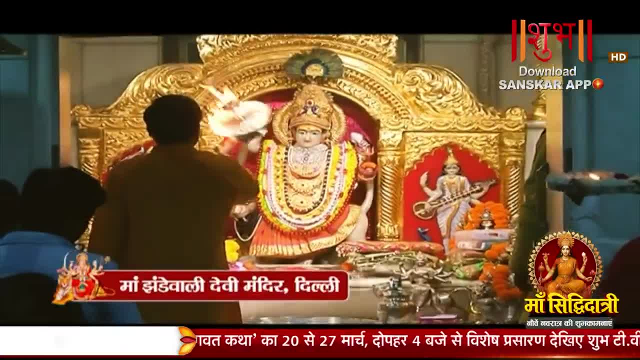 A man in a brown shirt places a garland around a statue of a goddess, her golden throne gleaming. Another smaller statue of a goddess playing a stringed instrument stands to her right.