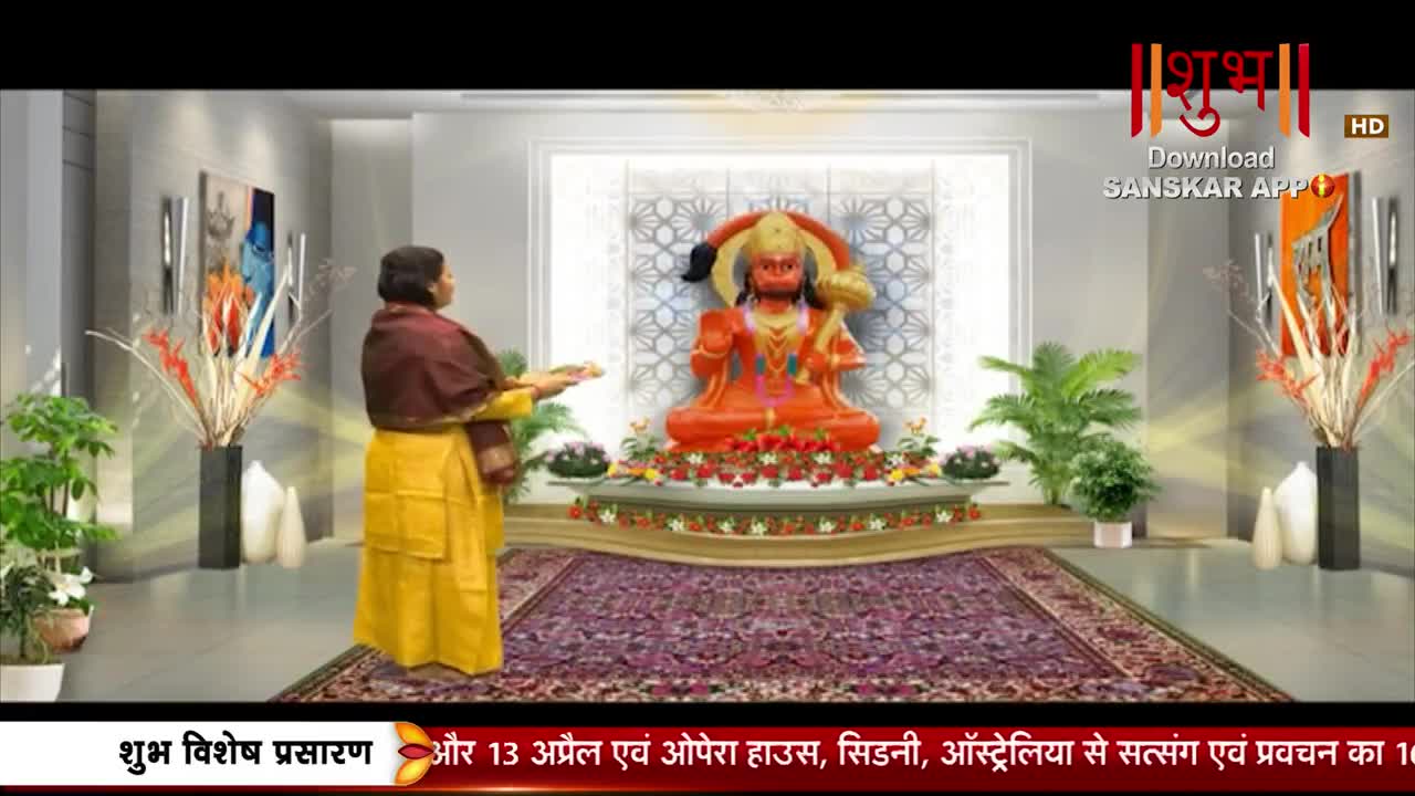 A woman in a yellow sari and maroon shawl holds a tray, facing a vibrant orange statue of Hanuman. The scene is set in a well-lit room, with potted plants and modern art on the walls, suggesting a television broadcast from Shubh TV.