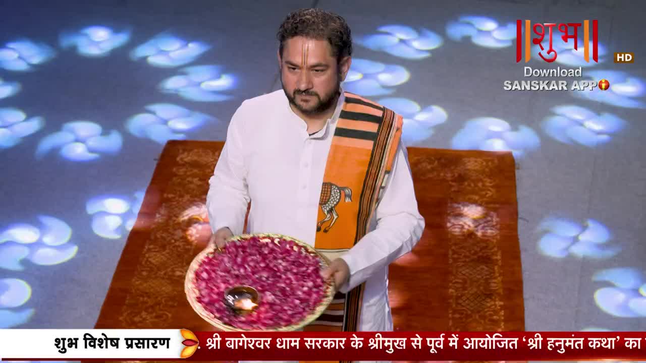 A man in white stands holding a woven basket overflowing with pink rose petals. A small, lit lamp rests in the center of the petals.