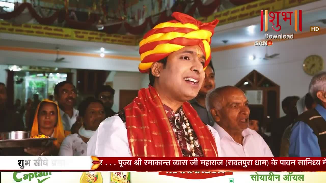 A man in a vibrant yellow and red turban sings, his face turned upwards. He's surrounded by a crowd of people, some watching intently, others looking elsewhere.