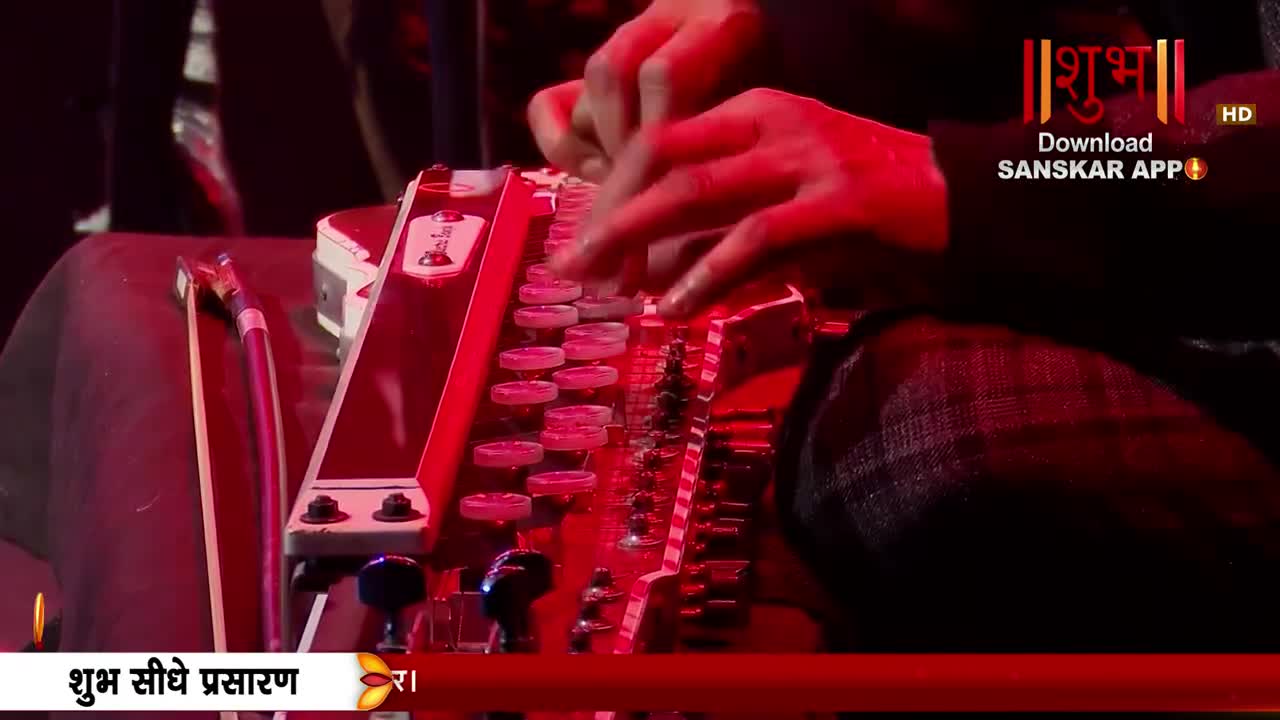 Fingers dance across the harmonium keys, a flurry of motion under the warm stage lights. The Shubh Sanskar App logo glows in the background, a reminder of the live broadcast from India.