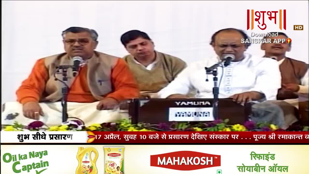 A man in an orange kurta and vest sings into a microphone, his hands resting on a harmonium. To his right, another man in white also sings, his gaze directed forward.