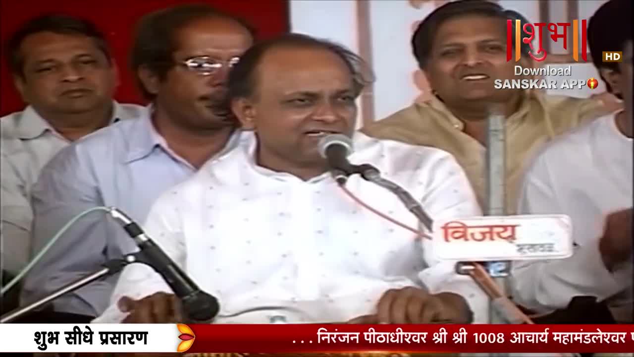 A man in a white shirt speaks into a microphone, his mouth open. Behind him, other men watch, some smiling. A banner at the bottom reads "Shubh Seedhe Prasaran" and another displays the Shubh TV logo.