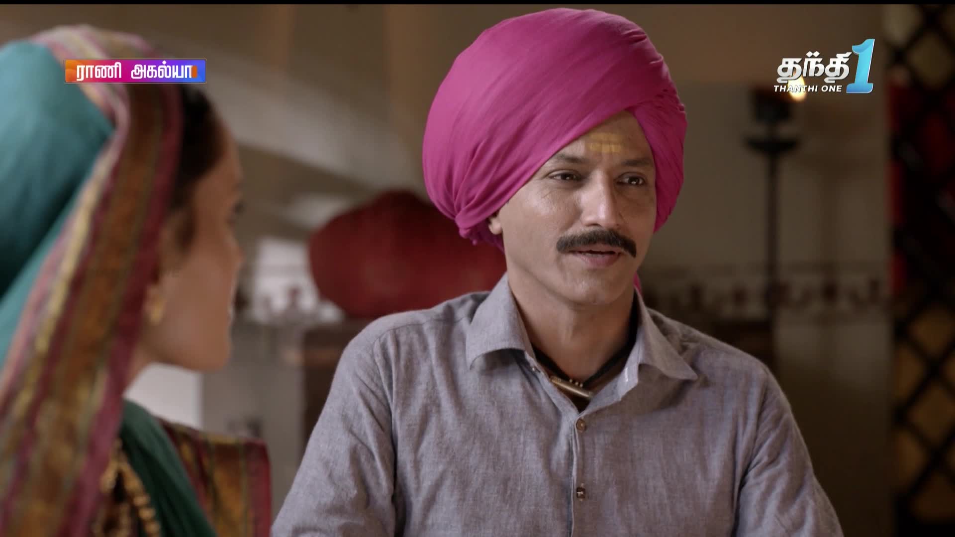 A man in a pink turban, wearing a grey shirt, is speaking, his face lit by a small dot of yellow on his forehead. Across from him, a woman in a green sari is listening, the scene likely unfolding on Thanthi One, as seen by the logo in the corner.
