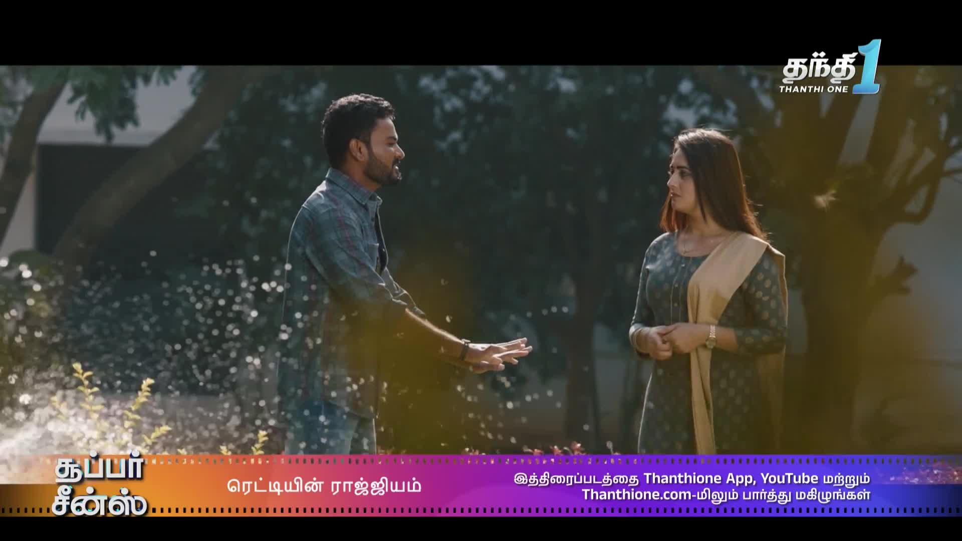A man gestures emphatically, his hands open, as he talks to a woman. Water droplets are suspended in the air around him, likely from a sprinkler, as the scene unfolds on Thanthi One.
