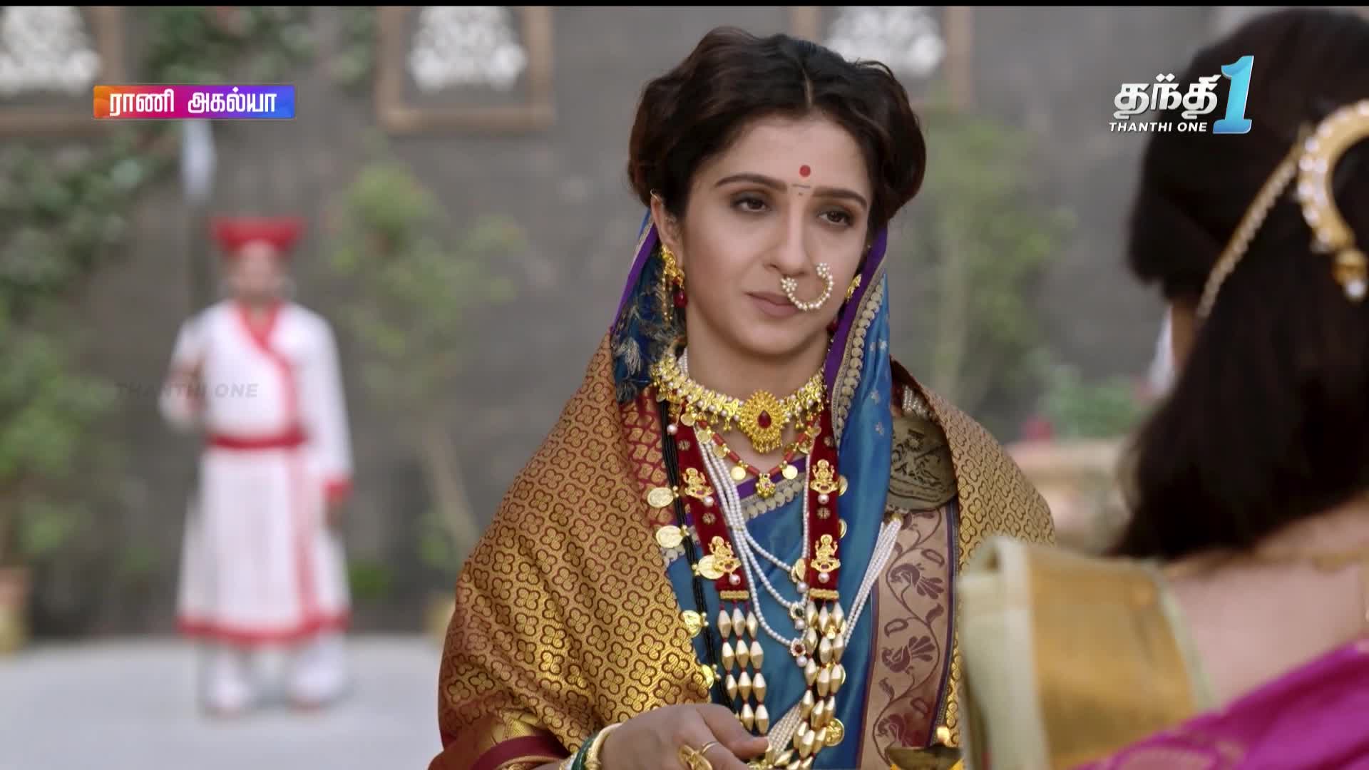 A woman, adorned in ornate jewelry and a blue sari, looks pensively toward someone out of frame. Behind her, a man in a white and red uniform stands in the distance, the scene likely from a Thanthi One broadcast.

