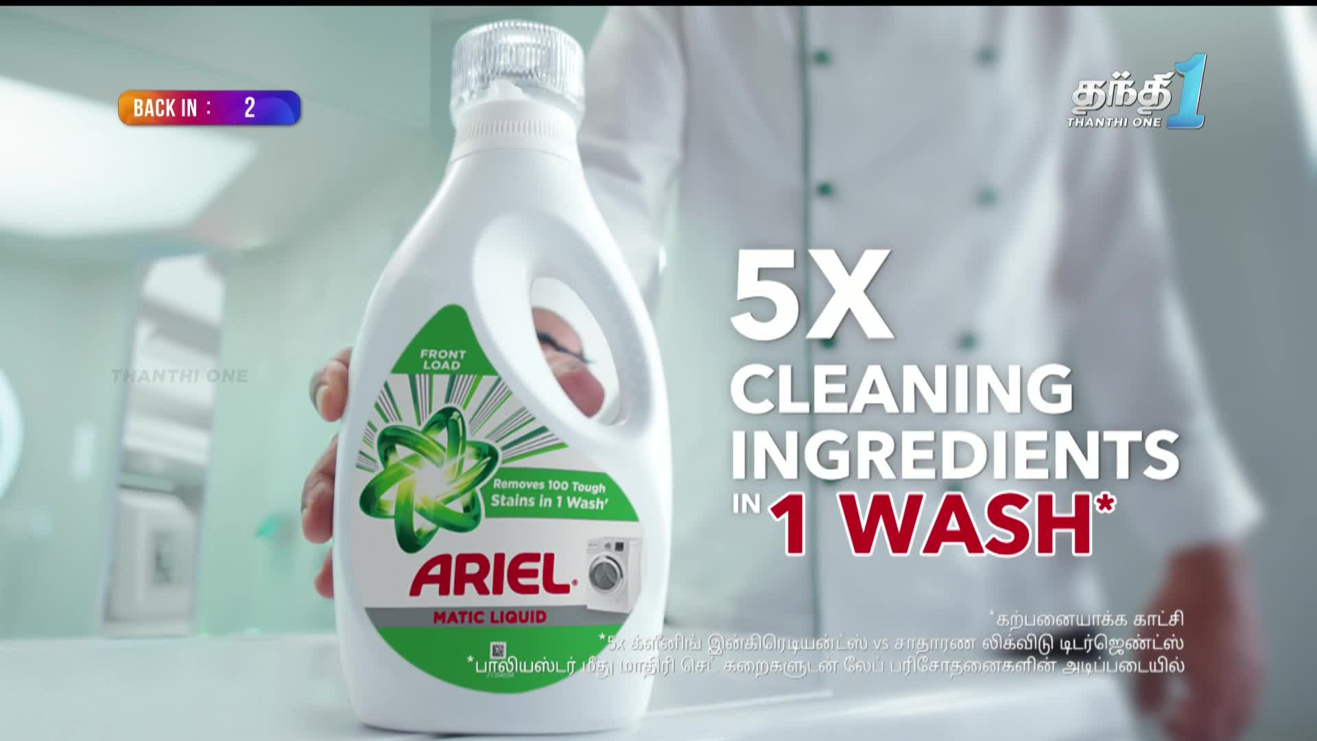 A hand holds a bottle of Ariel Matic Liquid, its label promising to remove stains. The Thanthi One logo is visible in the corner, and text advertises "5X cleaning ingredients in 1 wash."
