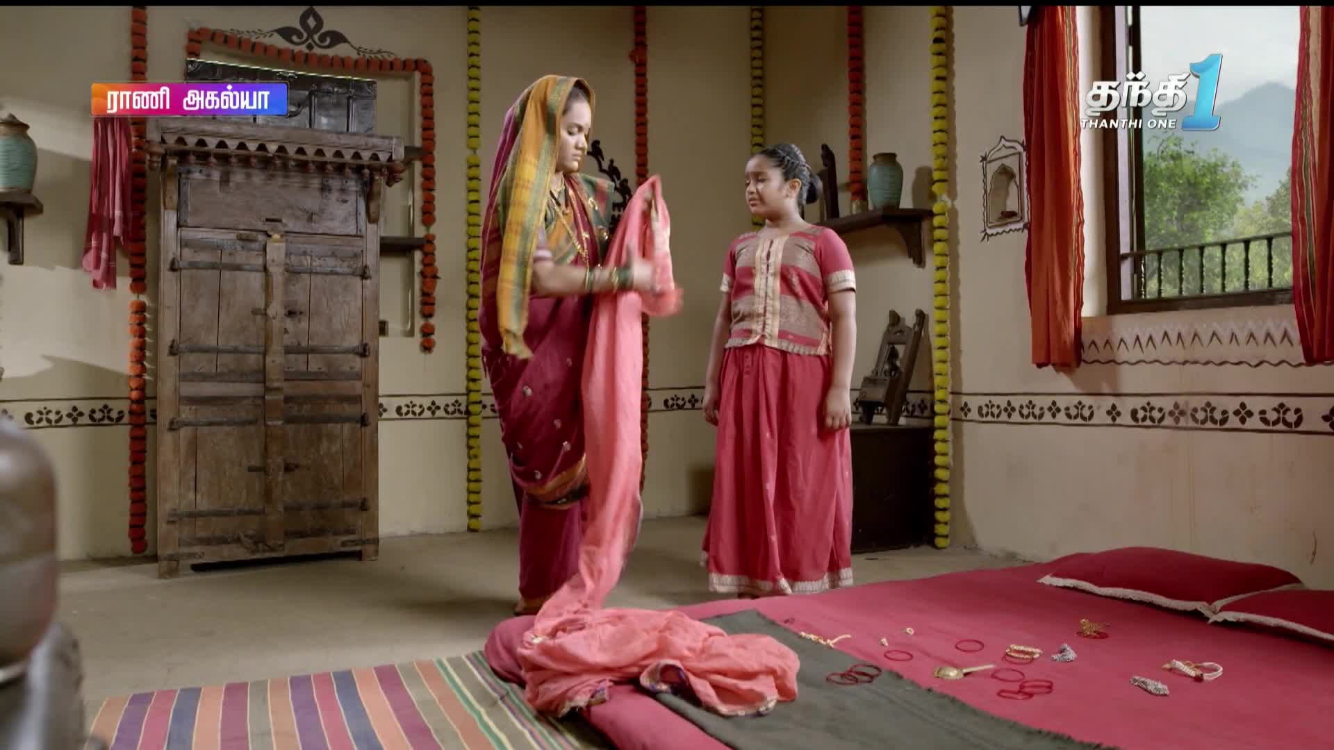 A woman in a maroon sari carefully folds a pink cloth in front of a young girl, with a bed and a large wooden cabinet behind them. The scene unfolds on Thanthi One, with the show's title displayed in the corner.
