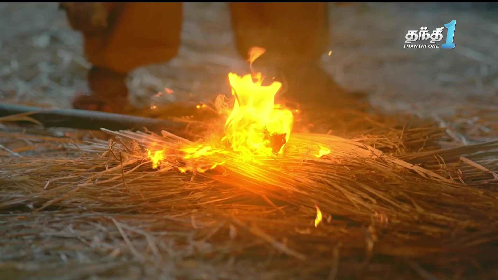 A small fire is burning brightly on a bed of dry straw, the flames dancing upwards.  Above, a person's legs are visible, and the Thanthi One logo is in the background.
