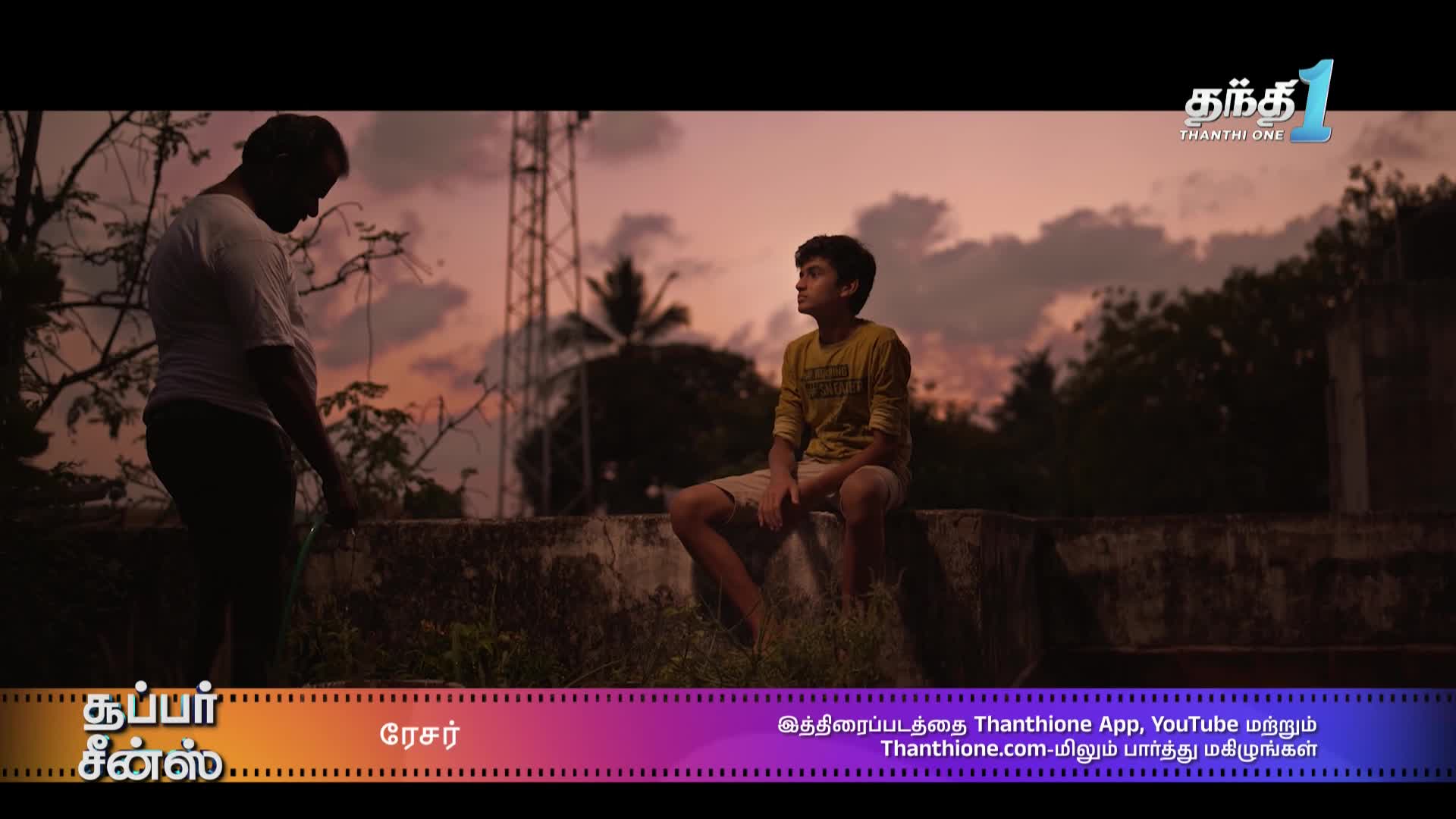 A young man sits on a low wall, gazing at the dusky sky. Nearby, a man in a white shirt tends to a hose, silhouetted against the fading light, perhaps in India.
