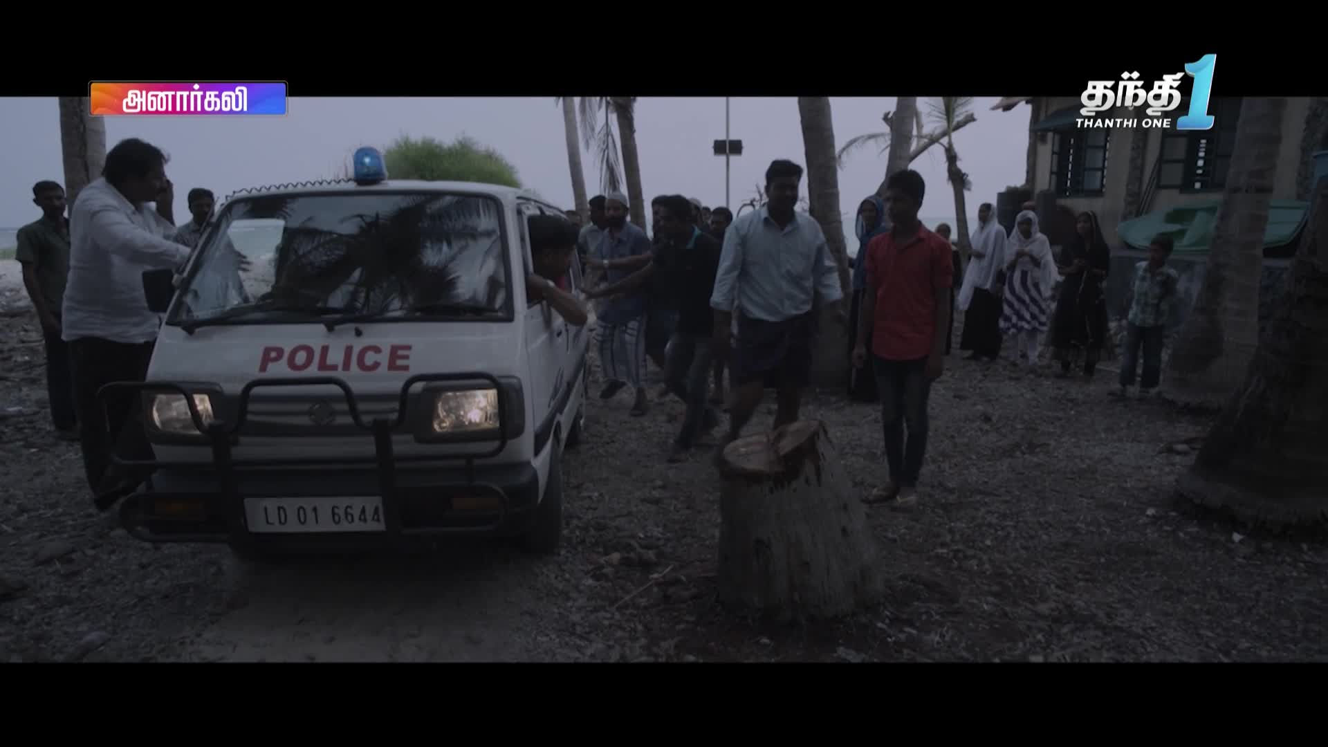 A white police van, its headlights on, is parked on a dirt road as people gather around. A group of people, some in traditional attire, are walking away from the van, likely the focus of a Thanthi One news report.
