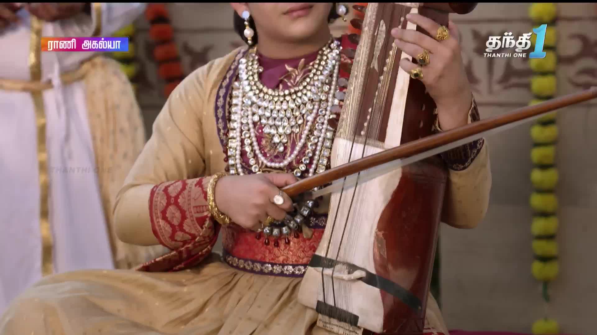 A musician, adorned in jewels and rich fabrics, draws the bow across the strings of a stringed instrument. The scene unfolds on Thanthi One, a television broadcast from India.
