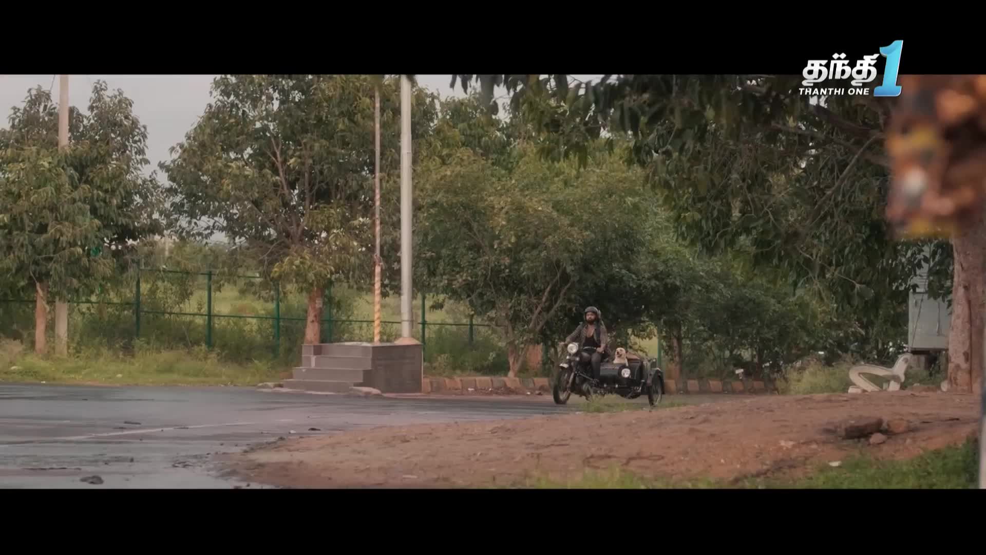 A motorcycle with a sidecar, likely from India, rolls along a dirt path. The scene is framed by trees and a cloudy sky, with the Thanthi One logo visible in the corner.
