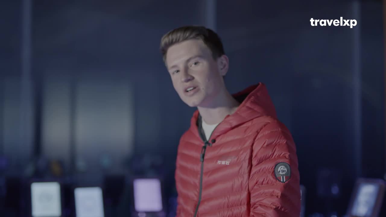 A young man in a red jacket speaks directly to the camera, a Travelxp logo visible in the corner. Behind him, the background is a dark, blurred space with what appear to be screens or monitors.
