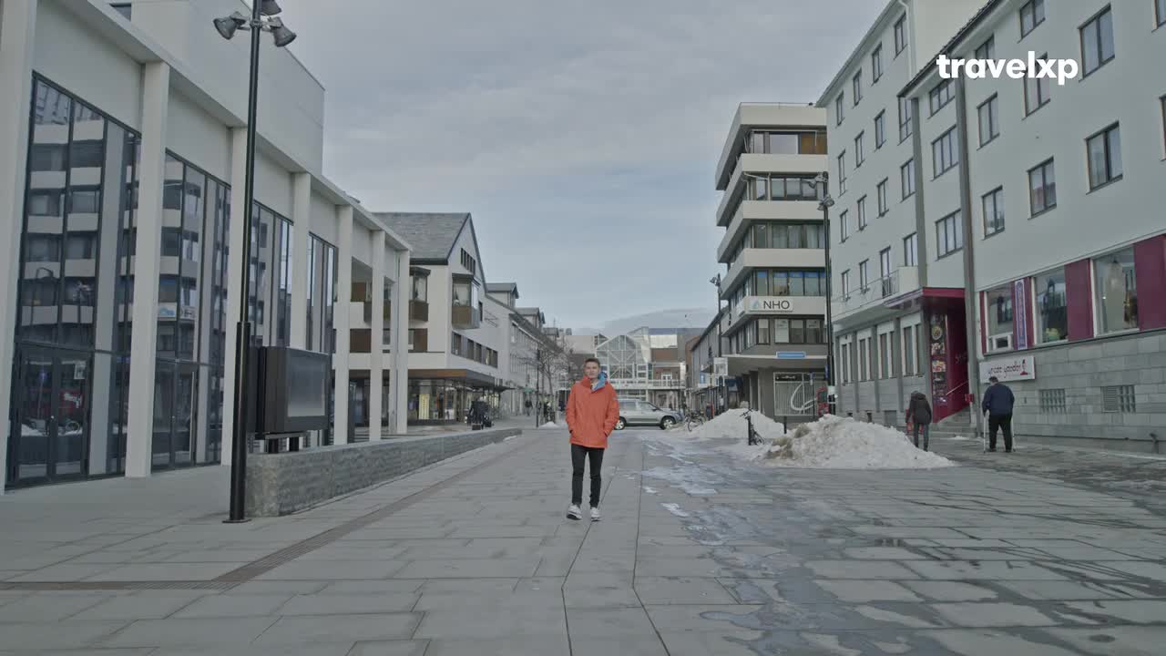 A man in an orange jacket walks towards me down a wide, paved street, flanked by modern buildings. Patches of snow remain on the sides, and the Travelxp logo sits in the top right.
