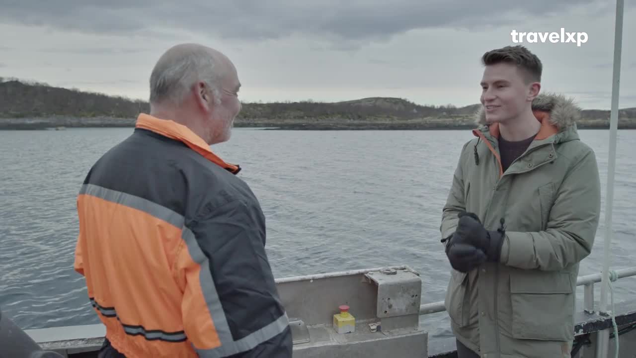The older man, wearing a bright orange vest, faces the water, while a younger man in a green jacket smiles, looking at him. They're on a boat, the sea stretching out behind them under a cloudy sky.
