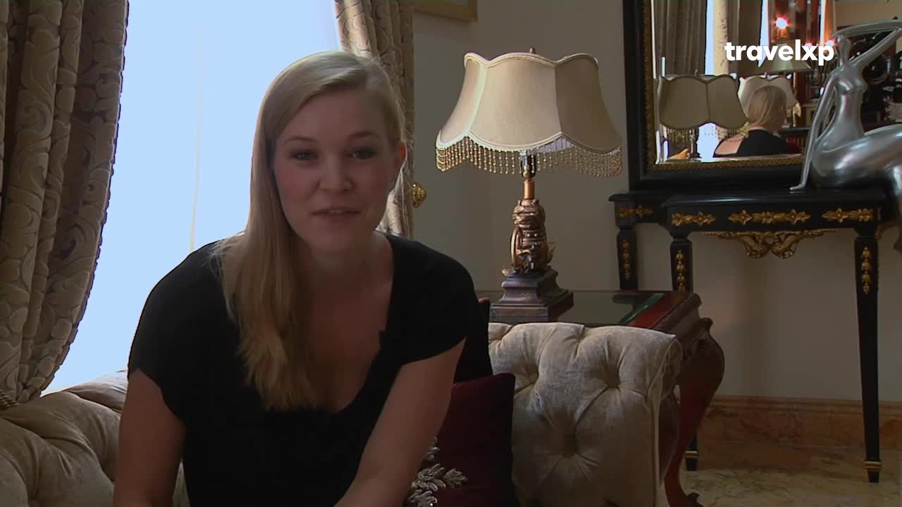 A woman with blonde hair sits on a plush couch, speaking directly to the camera. A Travelxp logo is visible in the upper right corner, and an ornate lamp sits behind her.
