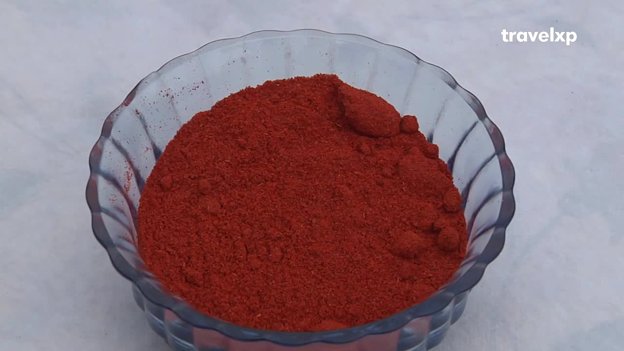 A clear glass bowl holds a generous mound of vibrant red powder. The Travelxp logo sits in the corner, suggesting this is part of a food presentation from India.
