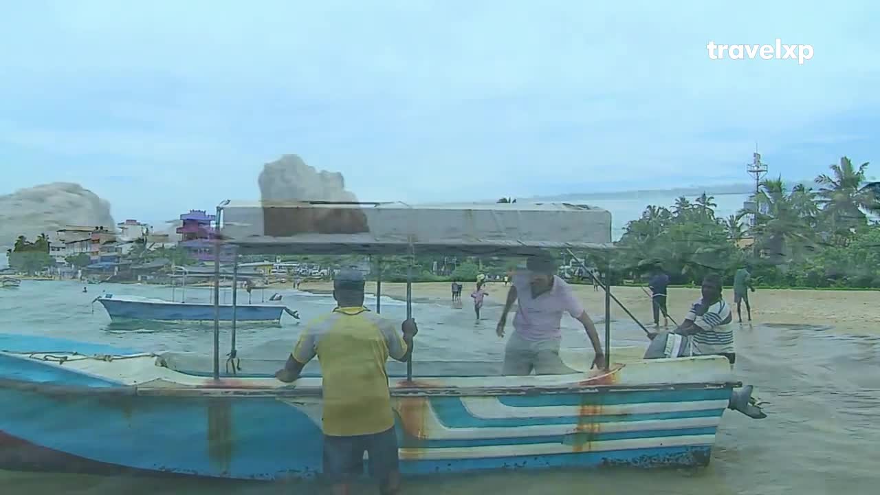 A man in a yellow shirt steadies the blue boat as another man steps out onto the wet sand. People wade in the shallows, while other boats bob in the water further out.
