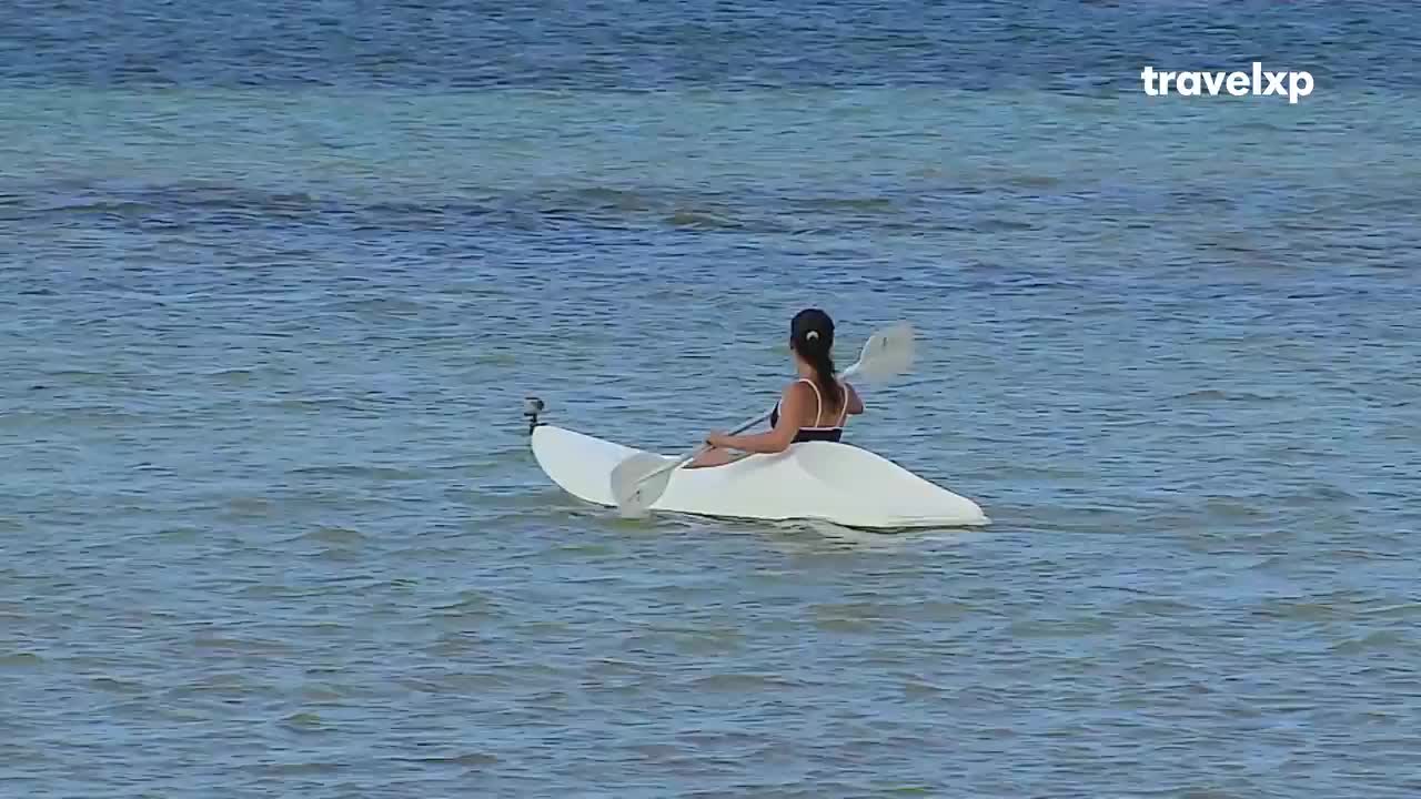 A woman sits in a white kayak, paddling through the calm, clear water. The camera angle is low, giving the impression of being right there on the water with her.
