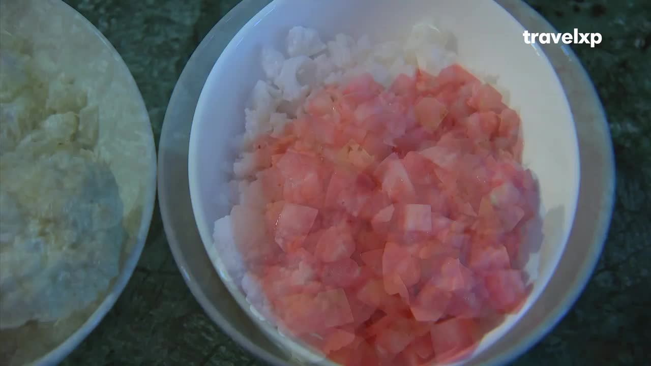 Two white bowls sit side-by-side, one filled with a pale, creamy substance, the other with a mix of white and diced pink tomatoes. The Travelxp logo is visible, suggesting a food-related segment from India.
