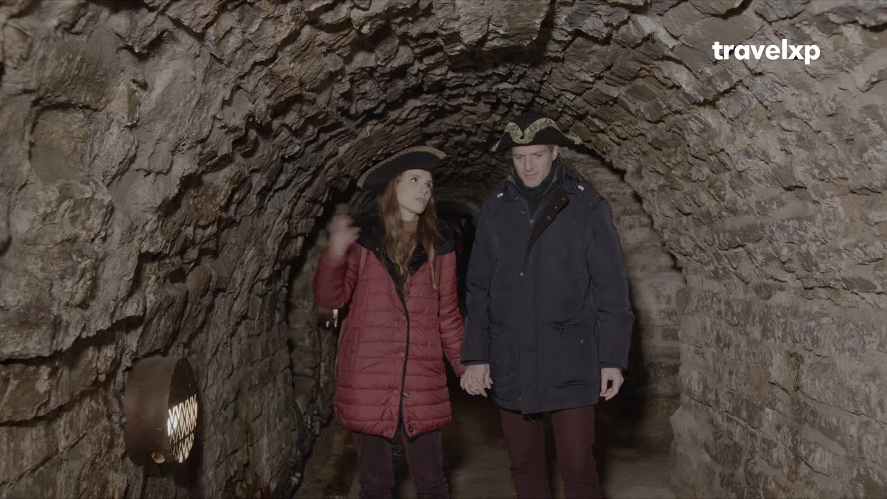 A couple, both wearing tricorn hats, stroll through a dimly lit, arched stone tunnel, holding hands. The woman gestures to the rough-hewn walls as they walk, while the man looks ahead.
