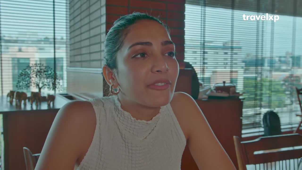 She's speaking, her mouth open as she looks toward someone off-camera. Sunlight streams through the blinds behind her, illuminating a cityscape visible through the windows.
