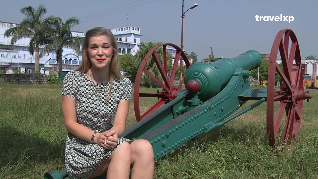 A woman in a patterned dress sits near a large, green cannon in front of a faded blue building. The Travelxp logo sits in the corner, suggesting this is a segment from a travel show in India.

