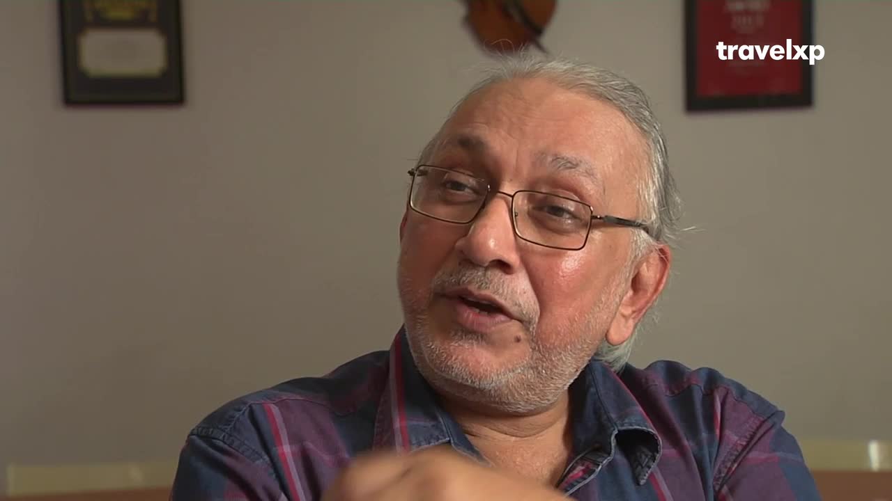 A man with glasses and a graying beard speaks, his mouth open in mid-sentence. The Travelxp logo is visible in the corner, suggesting a television interview.
