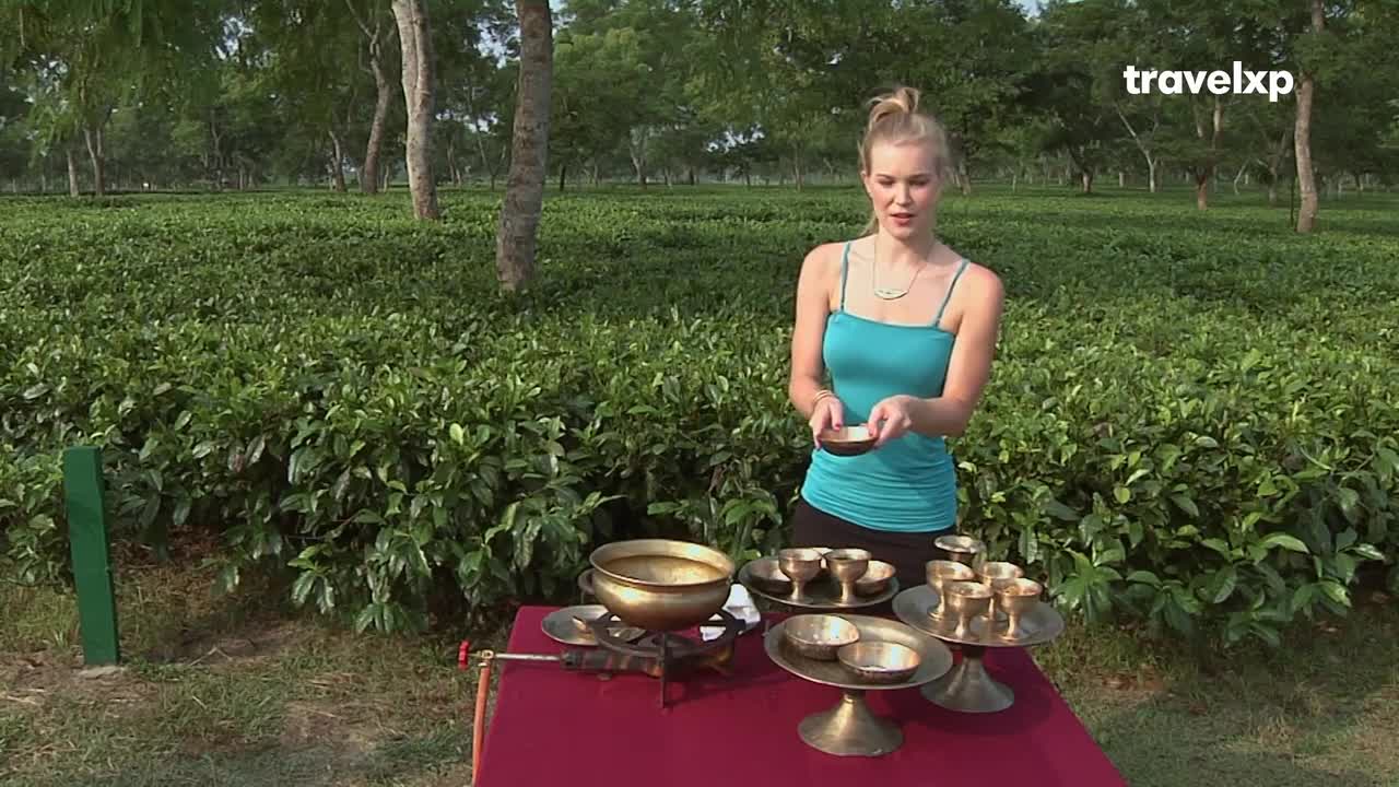 A woman in a teal top holds a small bowl, presenting it to the camera. Behind her, a tea plantation stretches out towards the trees, and a red table holds a collection of brass bowls and a burner.
