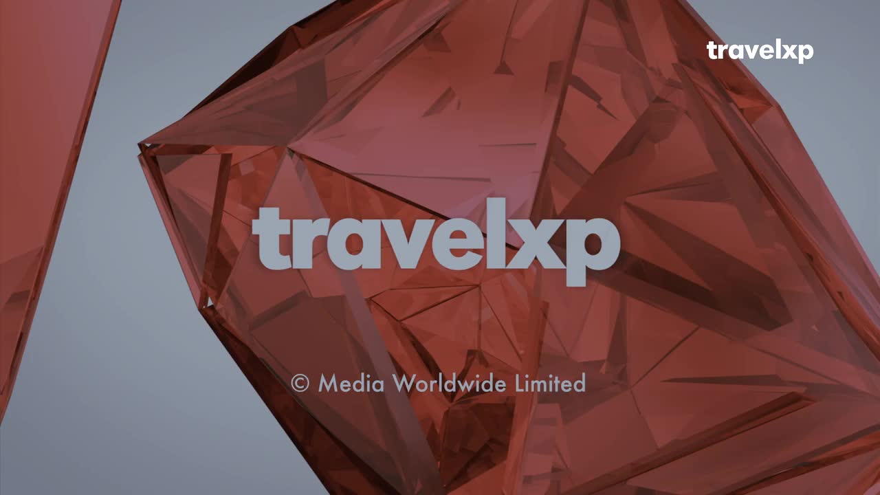 A translucent, ruby-colored geometric shape dominates the frame, with the "travelxp" logo overlaid in a stark, light gray. The logo appears against the backdrop of the abstract form, a visual element often associated with travel and exploration.
