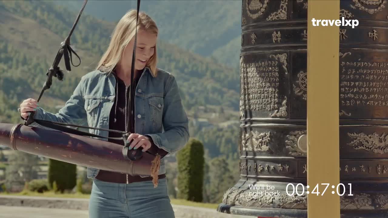 A woman in a denim jacket smiles, holding a long, cylindrical object. The camera focuses on her as she stands near a large, ornate bell, with mountains visible in the background.
