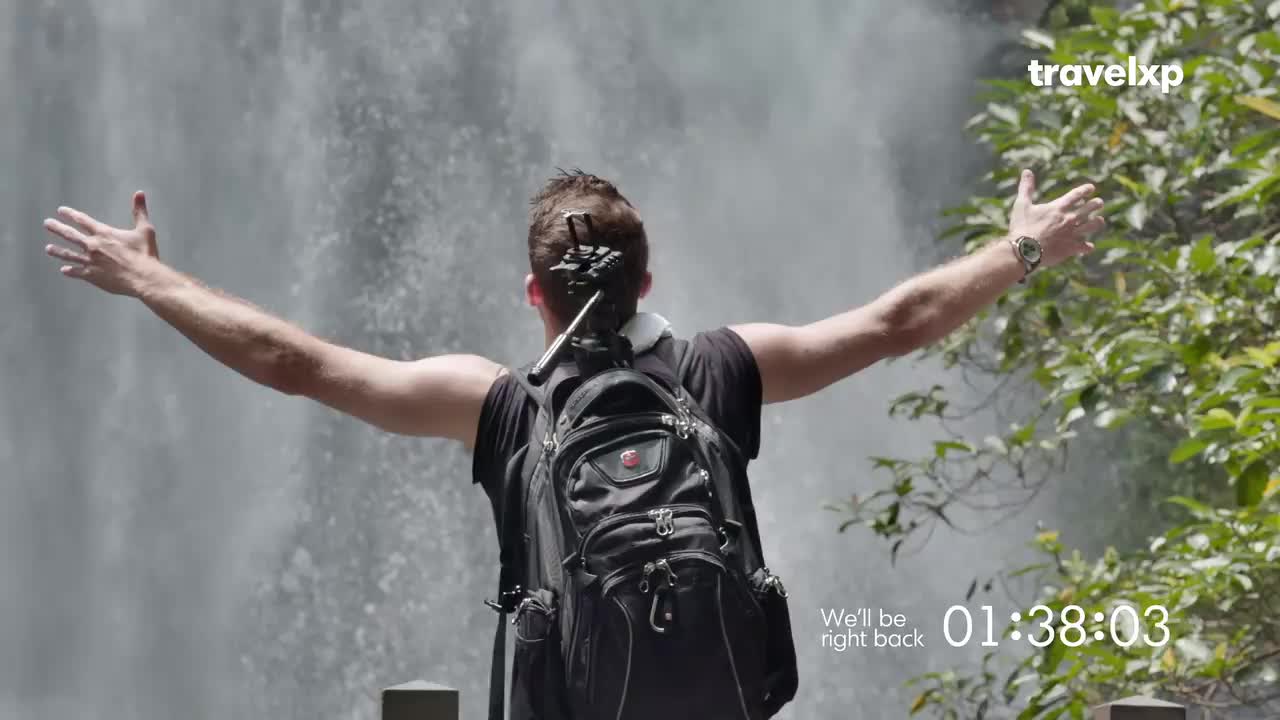 A man stands with his arms outstretched, facing a cascading waterfall. He's wearing a backpack and a small camera rig on his head, with a Travelxp logo in the corner.
