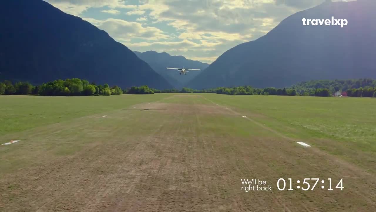 A small plane is descending towards a grassy airstrip, framed by towering mountains. The Travelxp logo sits in the corner, and a timer counts down until the show resumes.
