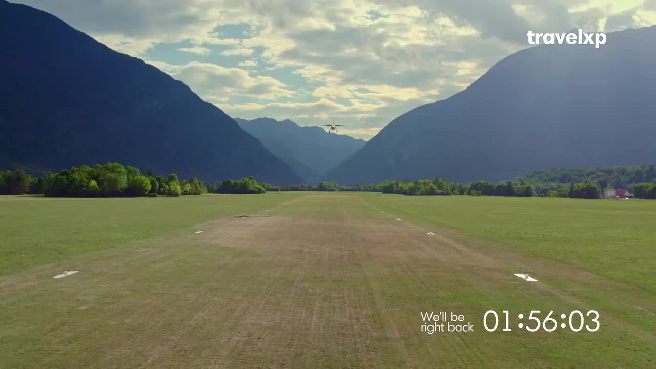 A small plane is coming in for a landing on a grassy runway, framed by towering mountains. The Travelxp logo and a "We'll be right back" message appear on the screen.
