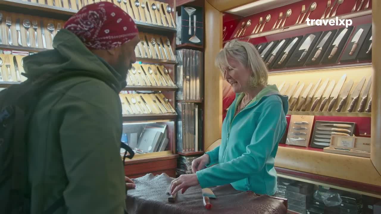 A woman in a turquoise jacket is arranging knives on a counter, smiling at a man wearing a red bandana. Behind them, shelves display rows of silverware and knives, likely in a shop.
