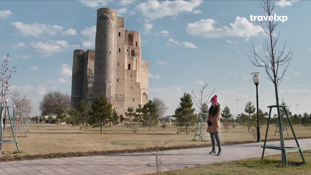 A woman in a red hat stands on a paved path, gazing at a towering, ancient structure. The sky is bright, and the Travelxp logo is visible in the corner.
