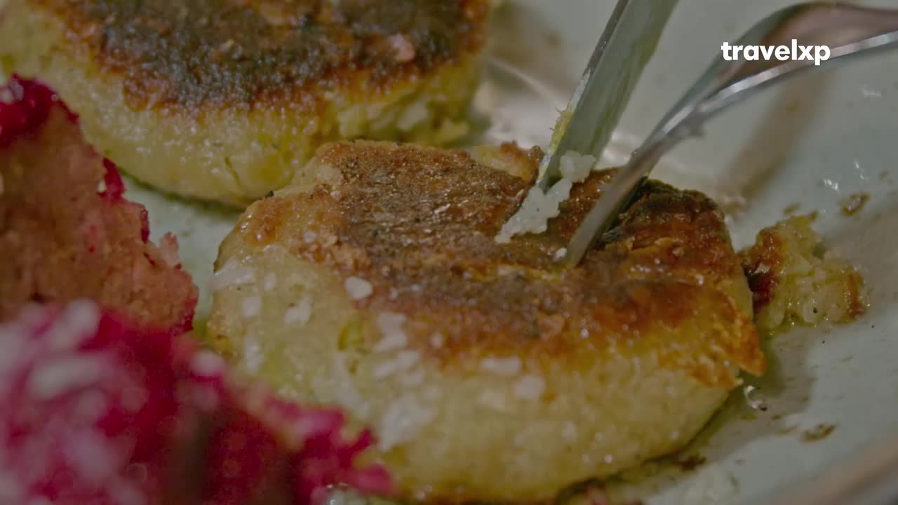 A knife and fork are slicing into a golden-brown patty, revealing its soft, fluffy interior. Beside it, a vibrant pink mound of what looks like chutney adds a splash of color to the plate.
