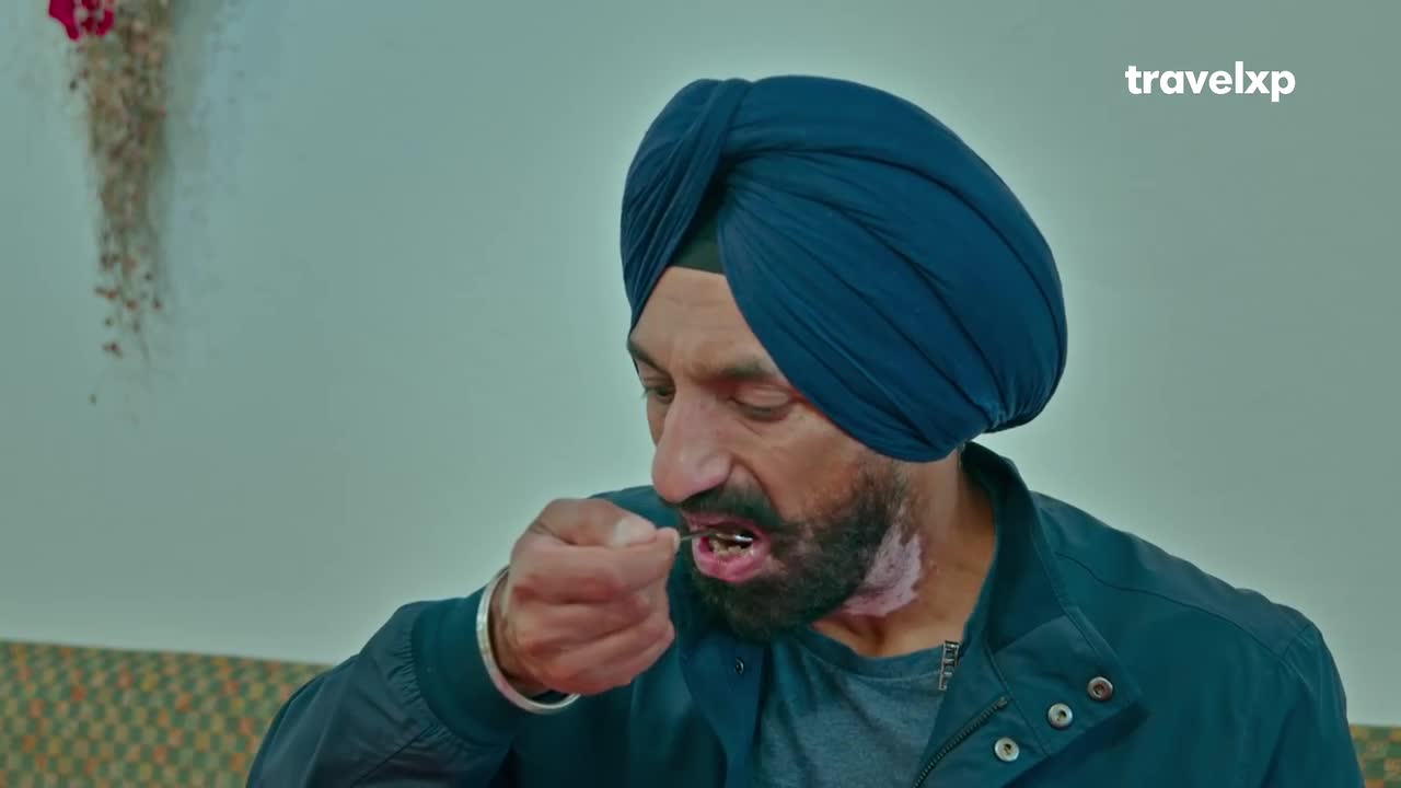 A man in a blue turban is bringing a small spoonful of food to his mouth. He's looking down at it, his face focused on the task.
