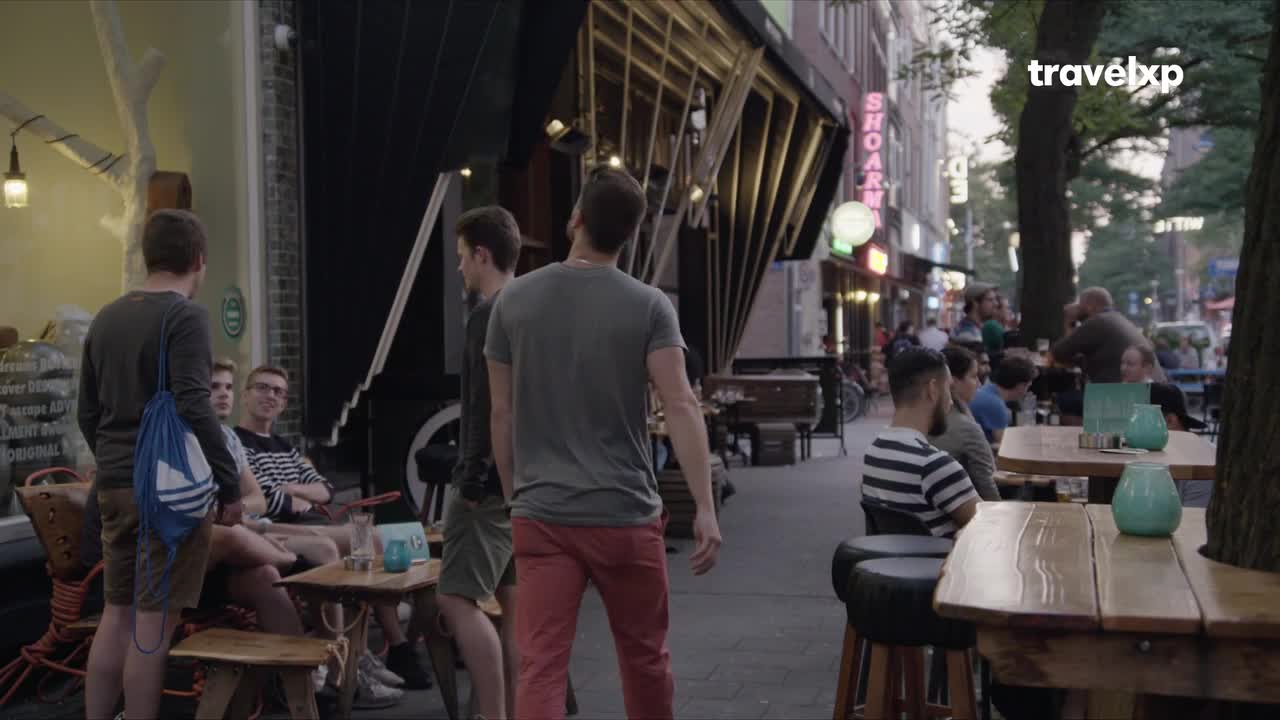 A group of young men mill around the sidewalk, some standing, others seated at outdoor tables. The neon sign of a restaurant glows in the background, and the Travelxp logo is visible in the upper right corner.
