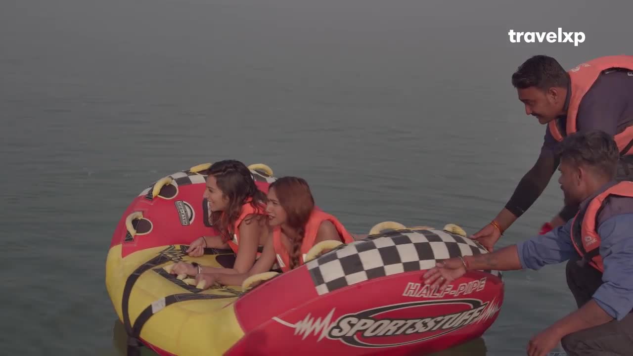 Two women in life vests are seated in a colorful inflatable, ready to be pulled across the water. Two men in matching vests are helping them, gently steadying the tube.

