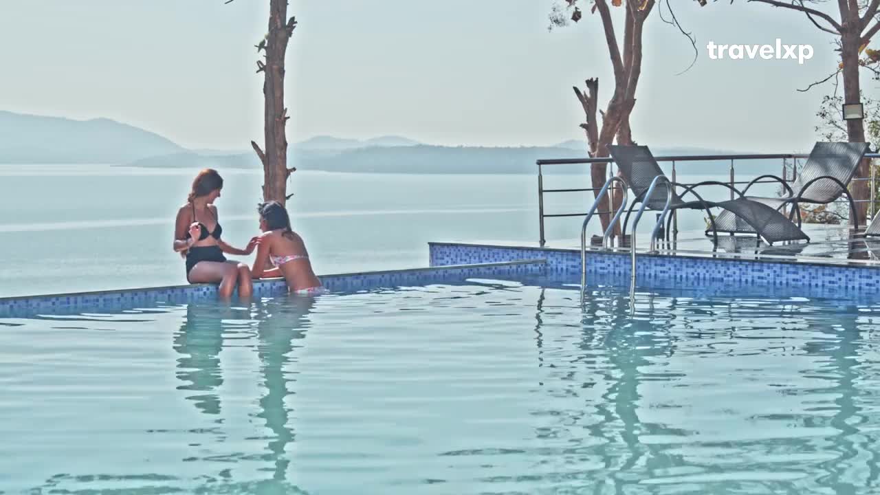Two women sit at the edge of a pool, legs dangling in the water. The backdrop is a vast, shimmering lake under a bright sky, and the scene feels peaceful.

