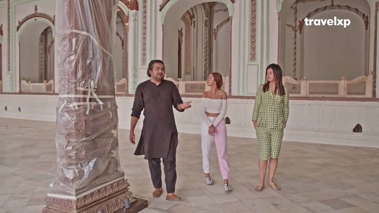 A man in a dark kurta gestures as he walks, leading two women through a large, ornate room. The Travelxp logo is in the corner, and a massive column wrapped in plastic stands in the foreground.
