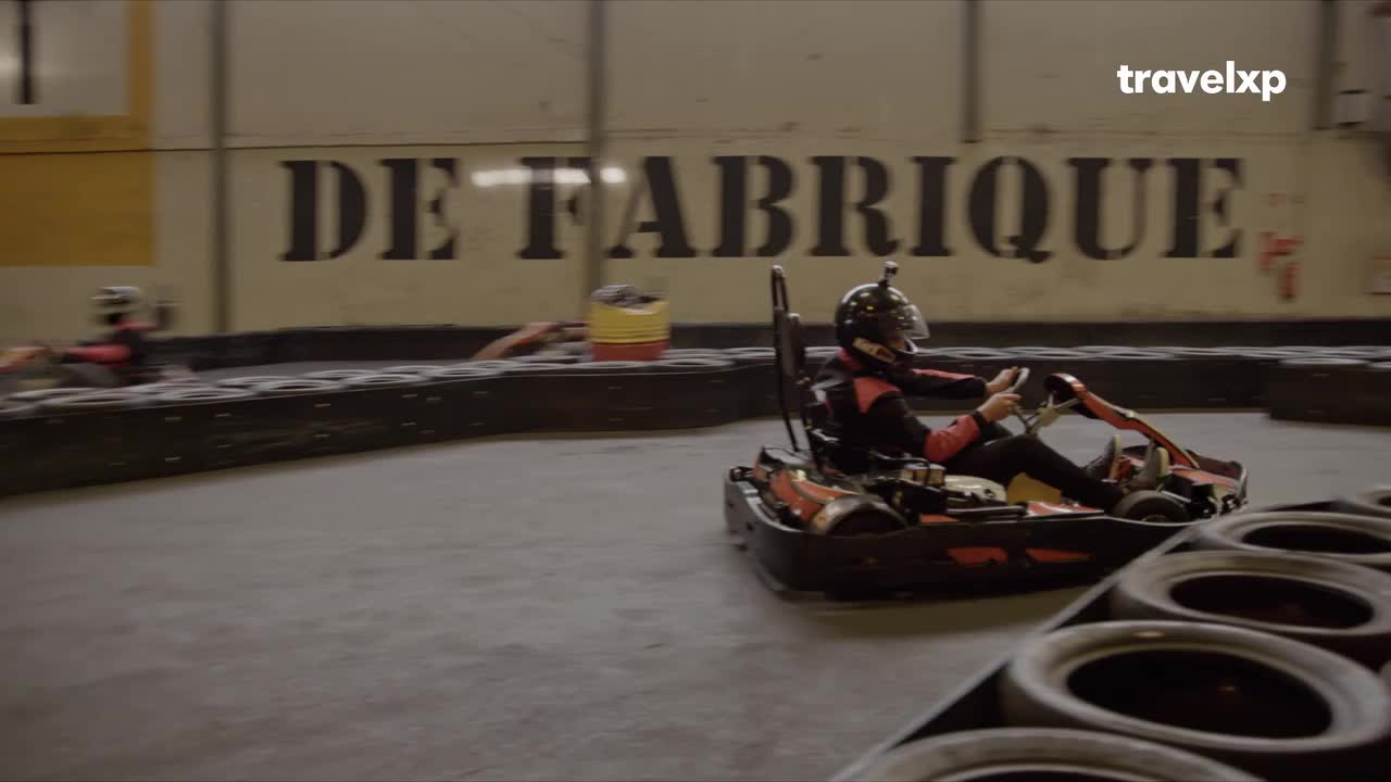 A go-kart, driven by someone in a helmet, whips around the track, the tires squealing as it navigates the turn.  Another kart is visible further back, also hugging the edge of the course.
