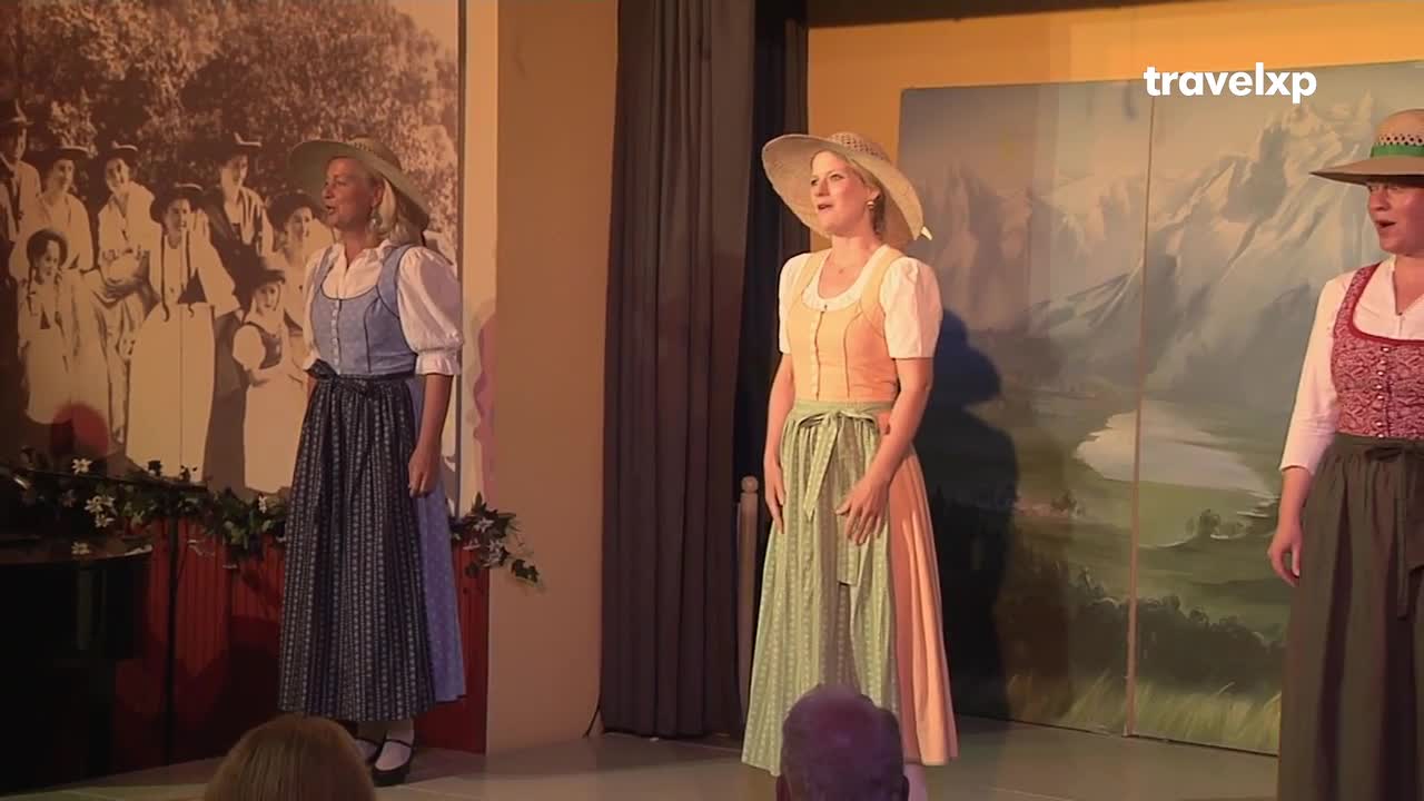 Three women in traditional dresses stand on a stage, singing with their mouths open. Behind them, a backdrop depicts mountains and a sepia-toned photo of a group of people.
